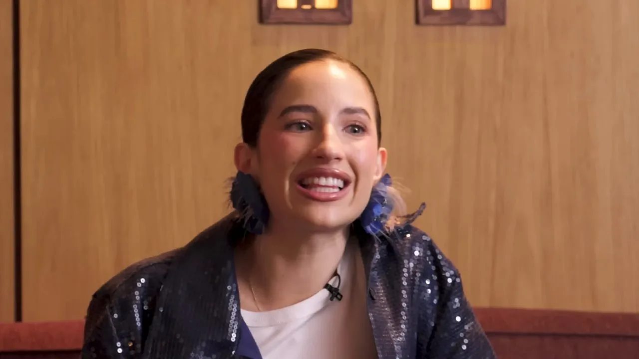 Aria Vega sonríe durante una entrevista en La Tobón Show; lleva el pelo recogido, aretes de plumas azules y una chaqueta de lentejuelas azul oscuro.